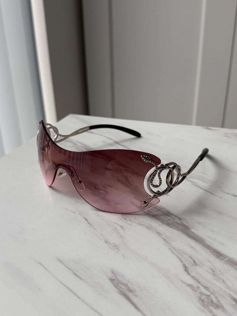 May include: Pink sunglasses with a silver frame and intricate detailing. The frame features a decorative design resembling a snake.