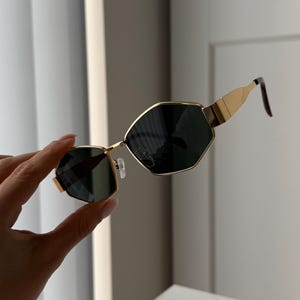 May include: Gold-toned sunglasses with a geometric, octagonal frame and dark lenses. The sunglasses are held in a hand.
