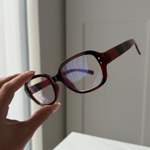 May include: A pair of eyeglasses with a dark red and black patterned frame. The lenses are clear with a slight blue tint. The glasses are held up by a hand against a white background.