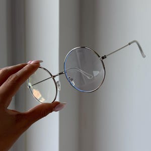 Może przedstawiać: Okrągłe okulary z metalową, srebrną oprawką i przezroczystymi soczewkami. Okulary mają cienką oprawkę i zauszniki, a trzyma je mostek nosowy. Soczewki mają lekkie niebieskie zabarwienie na krawędziach.