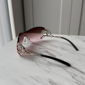 May include: A pair of pink sunglasses with a silver frame and black arms. The frame features a decorative design with interlocking circles.