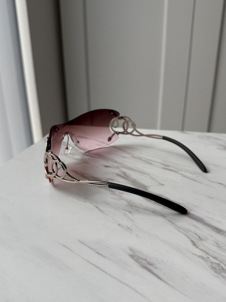 May include: A pair of pink sunglasses with a silver frame and black arms. The frame features a decorative design with interlocking circles.