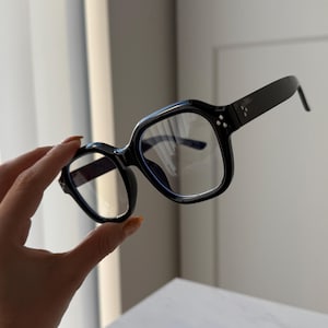 Könnte beinhalten: Schwarze Brille mit quadratischem Rahmen und klaren Gläsern. Die Brille hat einen dicken, glänzenden Rahmen mit drei kleinen silbernen Punkten an den Bügeln. Die Bügel sind gerade und erstrecken sich nach hinten.