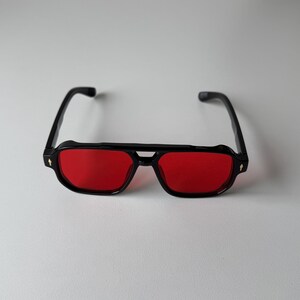 May include: Black sunglasses with red lenses. The glasses have a rectangular shape with a double bridge and a small decorative detail on the temples. The sunglasses are set against a white background.