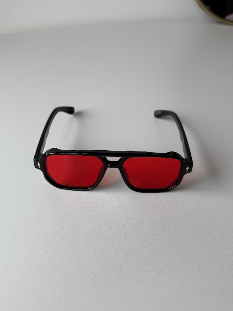 May include: Black sunglasses with red lenses. The glasses have a rectangular shape with a double bridge and a small decorative detail on the temples. The sunglasses are set against a white background.
