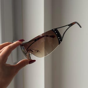May include: Brown gradient sunglasses with a black frame and decorative rhinestones. The sunglasses have a curved, shield-like design and a tortoiseshell pattern on the temples. The lenses transition from a darker shade at the top to a lighter shade at the bottom.