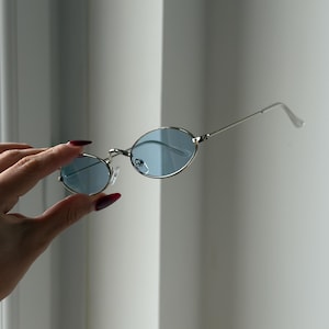 Könnte beinhalten: Ovale Sonnenbrille mit silbernem Rahmen und hellblauen Gläsern. Die Sonnenbrille wird vor einem weißen Hintergrund gehalten. Die Gläser sind hellblau und der Rahmen ist glänzend silber.