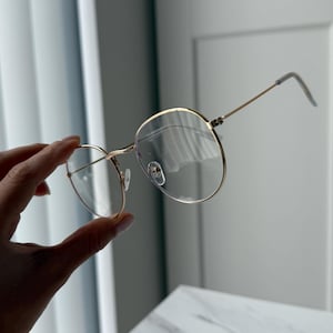 Puede incluir: Gafas redondas con una fina montura de metal dorado. Las lentes son transparentes y las patillas son de color dorado a juego. Las gafas están sujetas por una mano, mostrando el diseño y el estilo de las gafas.
