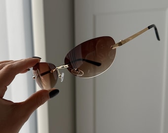 May include: Rimless sunglasses featuring oval-shaped brown gradient lenses. The eyewear has thin gold-tone metal temples and a matching gold-tone bridge. The temple tips are finished with black plastic covers, and the nose pads are clear.