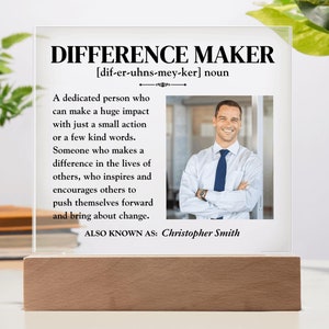 May include: A white acrylic plaque with a black border and text that reads "Difference Maker [dif-er-uhns-mey-ker] noun" and a definition of the word. The plaque also includes a photo of a man in a white shirt and blue tie. The text "ALSO KNOWN AS: Christopher Smith" is at the bottom of the plaque.