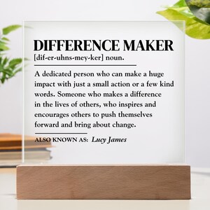 May include: A white acrylic sign with a black border and black text that reads "DIFFERENCE MAKER [dif-er-uhns-mey-ker] noun. A dedicated person who can make a huge impact with just a small action or a few kind words. Someone who makes a difference in the lives of others, who inspires and encourages others to push themselves forward and bring about change. ALSO KNOWN AS: Lucy James"
