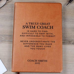 May include: A brown leather-bound notebook with the text "A Truly Great Swim Coach" and the name "Coach Smith" and the year "2025" embossed on the cover. The notebook has a ribbon bookmark. Glasses and a pen are on the wooden surface.