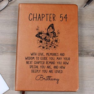 May include: A brown leather journal with the text "CHAPTER 54" and a butterfly design. The journal includes a personalized message and the name "Brittany". A bookmark is visible.