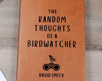 May include: A brown leather journal with the text "THE RANDOM THOUGHTS OF A BIRDWATCHER" in black. The journal also features a bird and binoculars graphic, and the name "DAVID SMITH". A bookmark is visible.