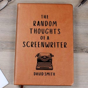 May include: A brown leather journal with the title "The Random Thoughts of a Screenwriter" and a typewriter graphic. The author's name, David Smith, is printed below. A bookmark is attached. Glasses and a pen are visible.