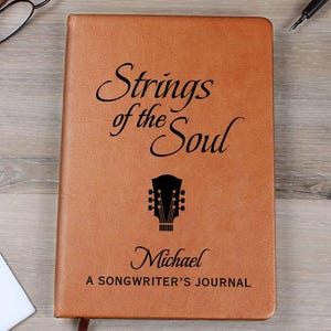 May include: A brown leather journal with the text "Strings of the Soul" and a guitar headstock graphic. The journal also includes the name "Michael" and the text "A Songwriter's Journal". A bookmark is visible.