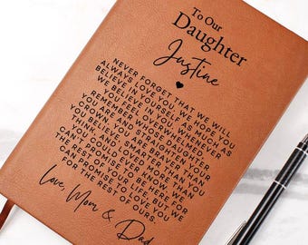 May include: A brown leather journal with the text "To Our Daughter Justine" and a heartfelt message. A black and silver pen rests beside the journal. The journal has a ribbon bookmark.