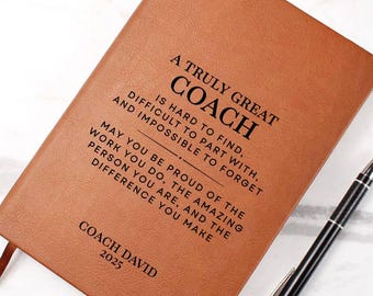 May include: A brown leather-bound journal with the text "A Truly Great Coach" and other inspirational text. A black and silver pen rests beside the journal. The journal includes a ribbon bookmark and the name "Coach David" and the year "2025".