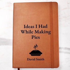 May include: A brown leather-bound journal with the text "Ideas I Had While Making Pies" and a black silhouette of a pie with steam rising from it. The name "David Smith" is printed below the pie.