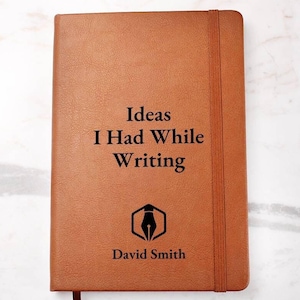 May include: A brown leather-bound journal with the text "Ideas I Had While Writing" and the name "David Smith" printed on the front cover. The journal has a black pen icon and a black elastic band.