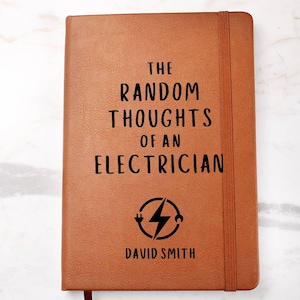 May include: A brown leather-bound notebook with the text "THE RANDOM THOUGHTS OF AN ELECTRICIAN" in black. Below the text is a graphic of a lightning bolt inside a circle, and the name "DAVID SMITH". The notebook has a brown elastic band and a ribbon bookmark.