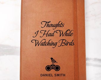 May include: A brown leather journal with the text "Thoughts I Had While Watching Birds" in black script. Below the text is a small bird perched on binoculars, and the name "DANIEL SMITH" is printed below. The journal has a ribbon bookmark.