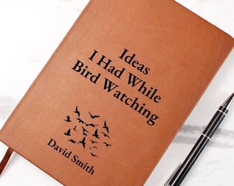 May include: A brown leather journal with the text "Ideas I Had While Bird Watching" and the name "David Smith" engraved on the front. A black pen is lying next to the journal.