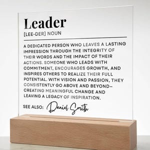 May include: A white acrylic plaque with a black wood base. The plaque has a definition of the word "Leader" with the text "See Also: Daniel Smith".