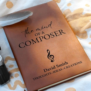 May include: A brown leather journal with the text "the mind of a COMPOSER" and a treble clef symbol. The journal also includes the name "David Smith" and the phrase "THOUGHTS.IDEAS.CREATIONS". A feather rests on the journal.