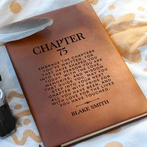 May include: A brown leather journal with the text "CHAPTER 75" at the top. The journal has an inspirational quote and the name "BLAKE SMITH" at the bottom. A feather rests on the journal.