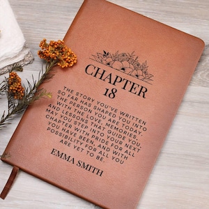 May include: A brown leather journal with the words "CHAPTER 18" and a floral design. The journal has a quote and the name "EMMA SMITH" printed on it. A sprig of orange flowers rests on the side.