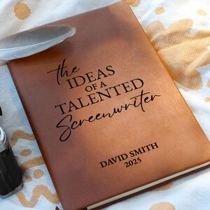 May include: A brown leather journal with the text "the IDEAS OF A TALENTED Screenwriter" and the name "DAVID SMITH 2025" printed on the cover. A feather rests on the journal. The journal is on a white and gold patterned surface.
