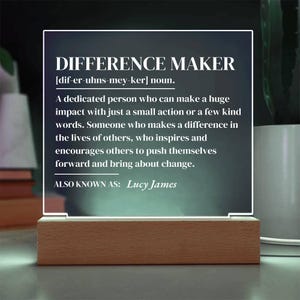 May include: A white acrylic sign with a wooden base, featuring the definition of a "Difference Maker". The text reads: "[dif-er-uhns-mey-ker] noun. A dedicated person who can make a huge impact with just a small action or a few kind words. Someone who makes a difference in the lives of others, who inspires and encourages others to push themselves forward and bring about change. ALSO KNOWN AS: Lucy James"