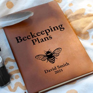 May include: A brown leather-bound notebook with the words "Beekeeping Plans" and a bee illustration. The name "David Smith" and the year "2025" are printed below the bee. A feather rests on the notebook.