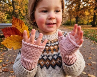 Op de afbeelding: Een kind draagt een lichtbeige gebreide muts en een crèmekleurige trui met een bruin, blauw en wit geometrisch patroon. Het kind draagt lichtroze vingerloze gebreide handschoenen en houdt verschillende feloranje en gele herfstbladeren vast.