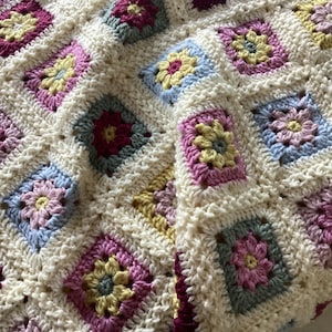 Könnte beinhalten: Eine handgehäkelte Decke mit einem wiederkehrenden Muster aus quadratischen Motiven. Jedes Quadrat enthält ein zentrales Blumenmuster in Gelb-, Rosa-, Lila- und Blautönen, die durch einen dicken, cremeweißen Garnrand verbunden sind.