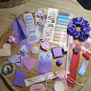 Könnte beinhalten: Eine Sammlung von lila und rosa Schreibwaren und Accessoires, darunter Lesezeichen, ein Scrunchie, Stifte, Aufkleber und eine kleine Kerze. Die Artikel sind auf einem Holztablett angeordnet.