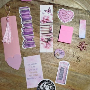 Op de afbeelding: Een verzameling roze en paarse briefpapierartikelen op een houten oppervlak. Inclusief bladwijzers, stickers, een pen en paperclips. De stickers bevatten boekenstapels, bloemmotieven en tekst. Een roze kwastjesbladwijzer en een hartvormige bladwijzer zijn ook aanwezig.