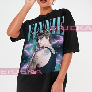 May include: Black oversized t-shirt featuring a graphic of a person with tattoos, set against a cosmic background with the name "VINNIE" in teal and white. The shirt is a unisex style.