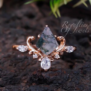 Peut inclure: Une bague en or rose avec une pierre de mousse agate et un design délicat et orné. La bague présente une grande pierre de mousse agate facetée en forme de diamant, entourée de petites pierres claires. La bande est fine et présente un motif floral.