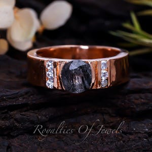 May include: A rose gold ring featuring a black gemstone and small clear gemstones on the band. The ring is resting on a dark brown wooden surface.