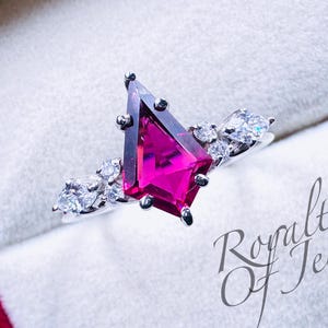 Peut inclure: Une bague en argent avec une grande pierre précieuse rose facetée de forme triangulaire. La bague est sertie de petites pierres précieuses claires autour de la pierre principale. La bague est présentée dans un écrin blanc avec le texte "Royalties of Jewel" imprimé en bas à droite.