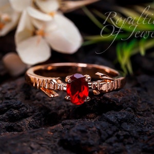 May include: A rose gold ring with a red oval-shaped gemstone set in a delicate arrow design. The ring is on a dark brown wooden surface.