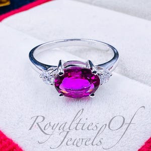 May include: A silver ring with a large oval-shaped pink gemstone surrounded by two smaller clear gemstones. The ring is in a red velvet ring box.