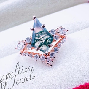 Könnte beinhalten: Ein roségoldener Ring mit einem großen, rautenförmigen Moosachatstein. Der Stein hat grüne und weiße Einschlüsse. Der Ring ist von kleineren, klaren Edelsteinen umgeben. Der Text "Royalties Of Jewels" ist sichtbar.