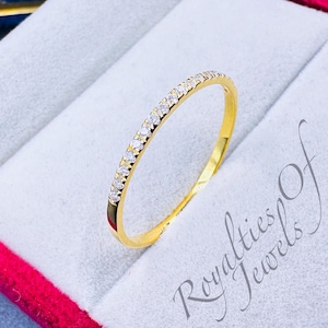 Pode incluir: Uma fina banda de ouro com uma fileira de pequenas pedras transparentes engastadas no metal. O anel está em uma caixa de veludo vermelho com as palavras "Royalties of Jewels" impressas na parte inferior.