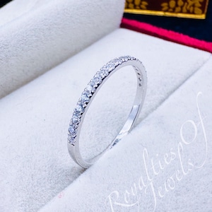 Pode incluir: Uma fina banda de prata com uma fileira de pequenas pedras preciosas transparentes engastadas em pavé. O anel é apresentado em uma caixa branca com forro vermelho e o texto "Royalties of Jewels" impresso no canto inferior direito.