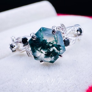 May include: A silver ring featuring a hexagonal moss agate gemstone with green and white patterns. The ring has a floral design with black accents on the band. The gemstone is the focal point, showcasing its unique natural inclusions.