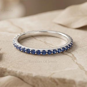 May include: A silver ring with a band of small, round, blue gemstones. The ring is set on a light-colored, textured surface, with blurred leaves in the background. The ring is a piece of jewelry.