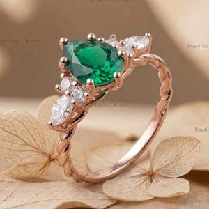 May include: A rose gold ring featuring a pear-shaped emerald center stone, accented by smaller white gemstones. The ring has a twisted band design and is displayed on a neutral-toned background, showcasing the jewelry's elegant details.